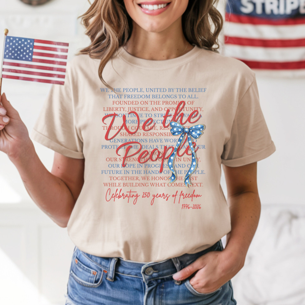 a soft beige unisex Gildan t-shirt with a crew neckline, short sleeves, layered patriotic front wording, a star-spangled bow graphic, a 1776-2026 commemorative detail, and an easy year-round fit
