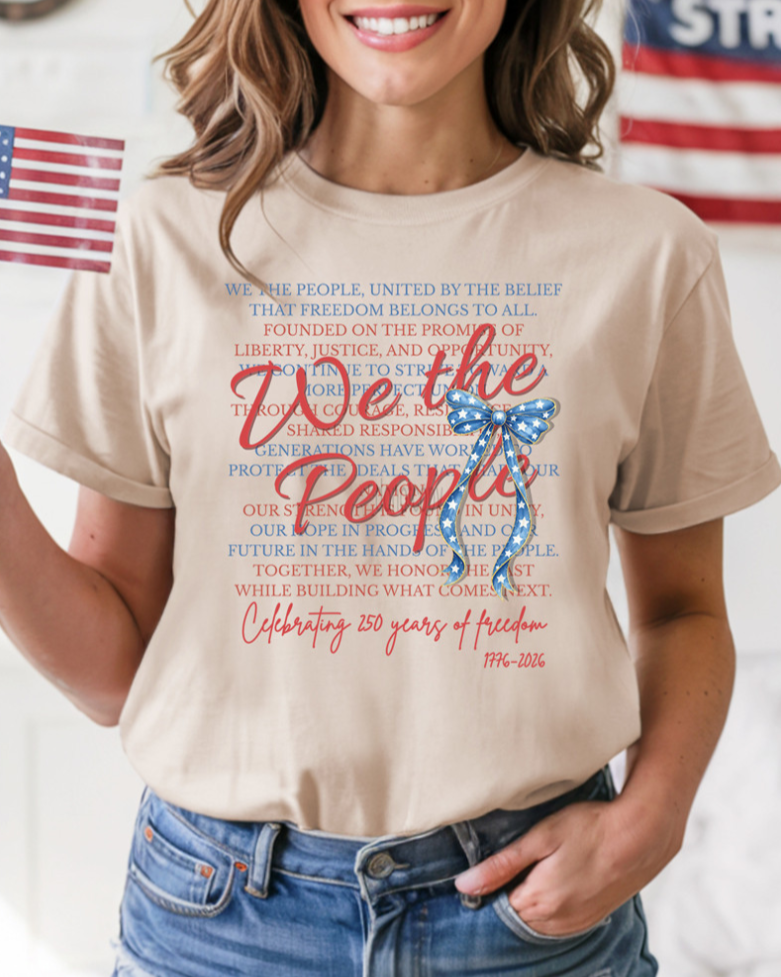 a soft beige unisex Gildan t-shirt with a crew neckline, short sleeves, layered patriotic front wording, a star-spangled bow graphic, a 1776-2026 commemorative detail, and an easy year-round fit