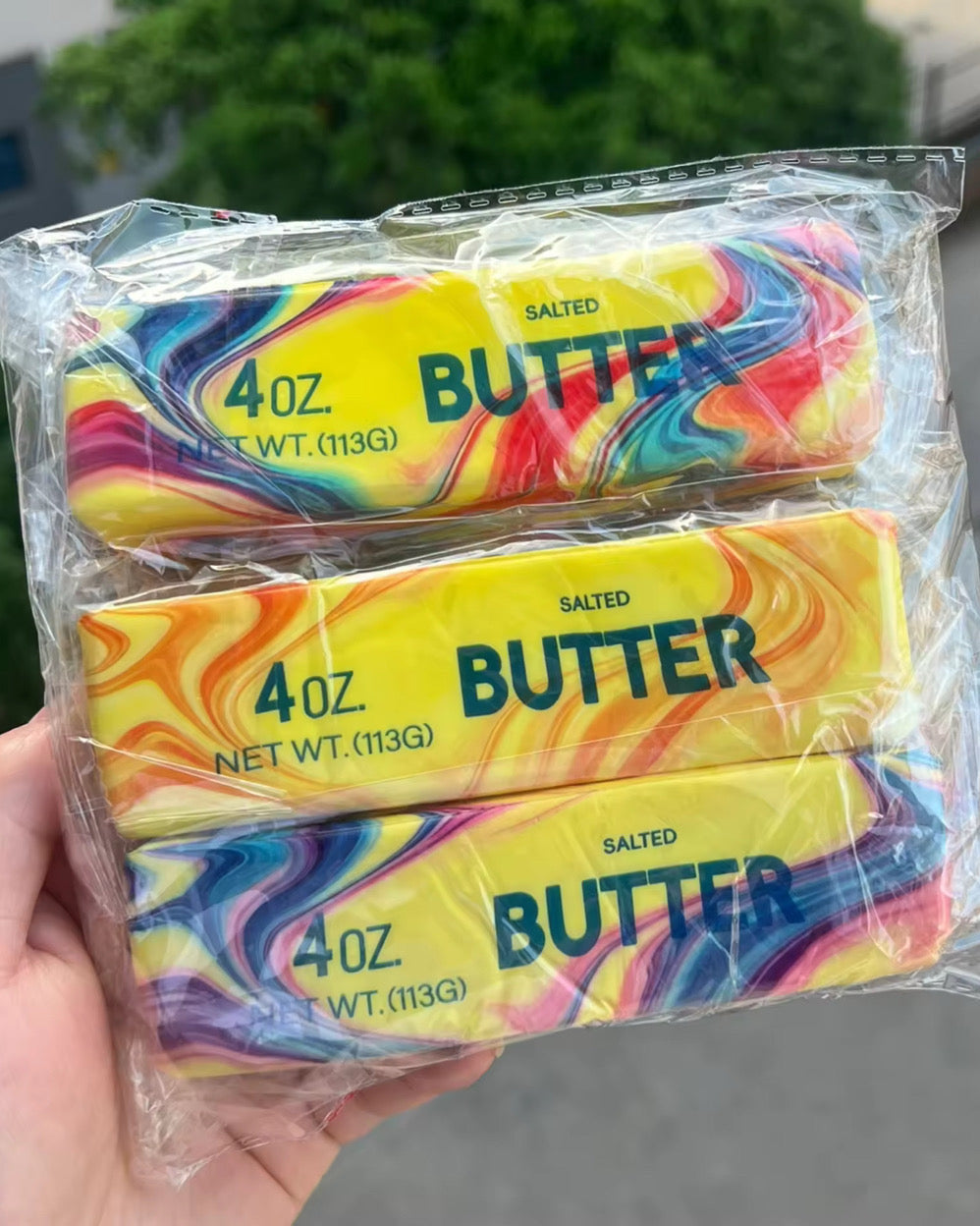 a 4oz butter-stick inspired squishy with a marbled design, salted butter label, soft squeezable texture, and clear individual packaging in colors from top to bottom rainbow swirl (red), citrus swirl (orange), and cotton candy swirl (pink)