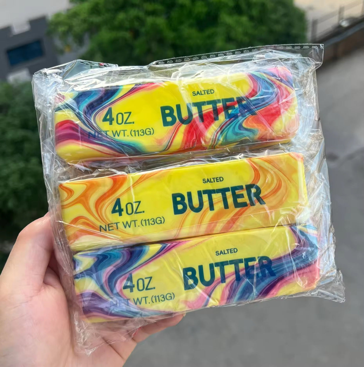 a 4oz butter-stick inspired squishy with a marbled design, salted butter label, soft squeezable texture, and clear individual packaging in colors from top to bottom rainbow swirl (red), citrus swirl (orange), and cotton candy swirl (pink)
