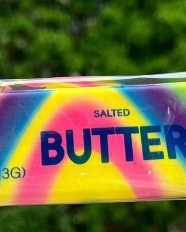 Packaged salted butter bar with colorful tie-dye design held by a hand, blurred greenery in the background.