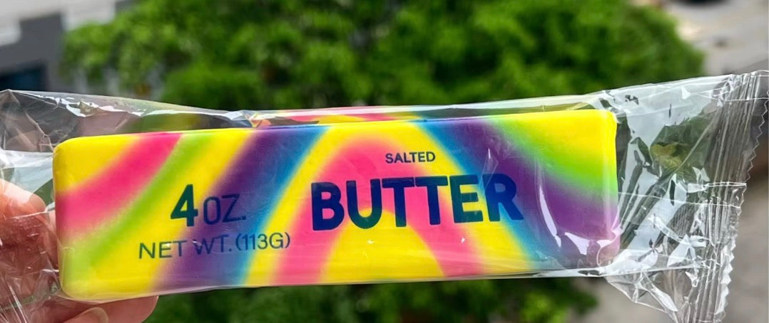 Packaged salted butter bar with colorful tie-dye design held by a hand, blurred greenery in the background.
