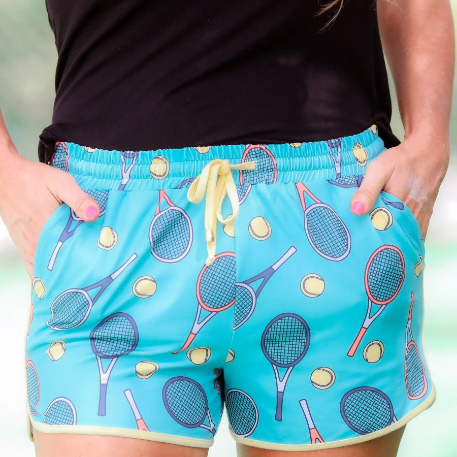 mid-rise relaxed fit activewear shorts with side pockets and a tie waist drawstring with gold bead accents, designed on a tennis pattern print fabric, cadet blue base, and yellow trim