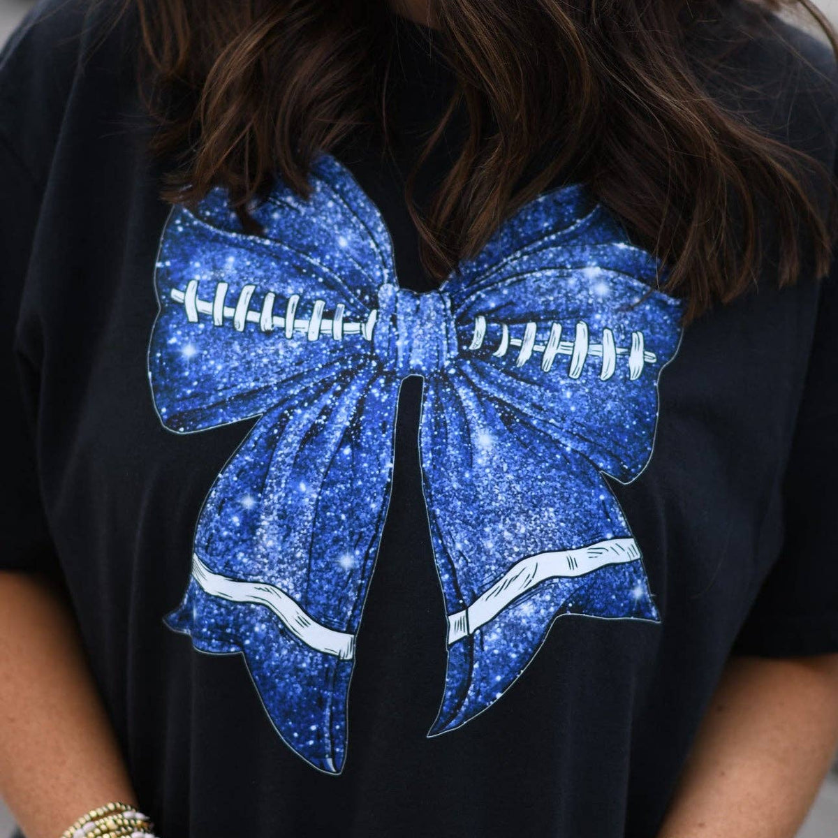 a black Comfort Colors tee crafted on soft garment-dyed cotton with a DTF-printed blue faux glitter football bow graphic, crew neckline, and short sleeves in both youth and adult sizing