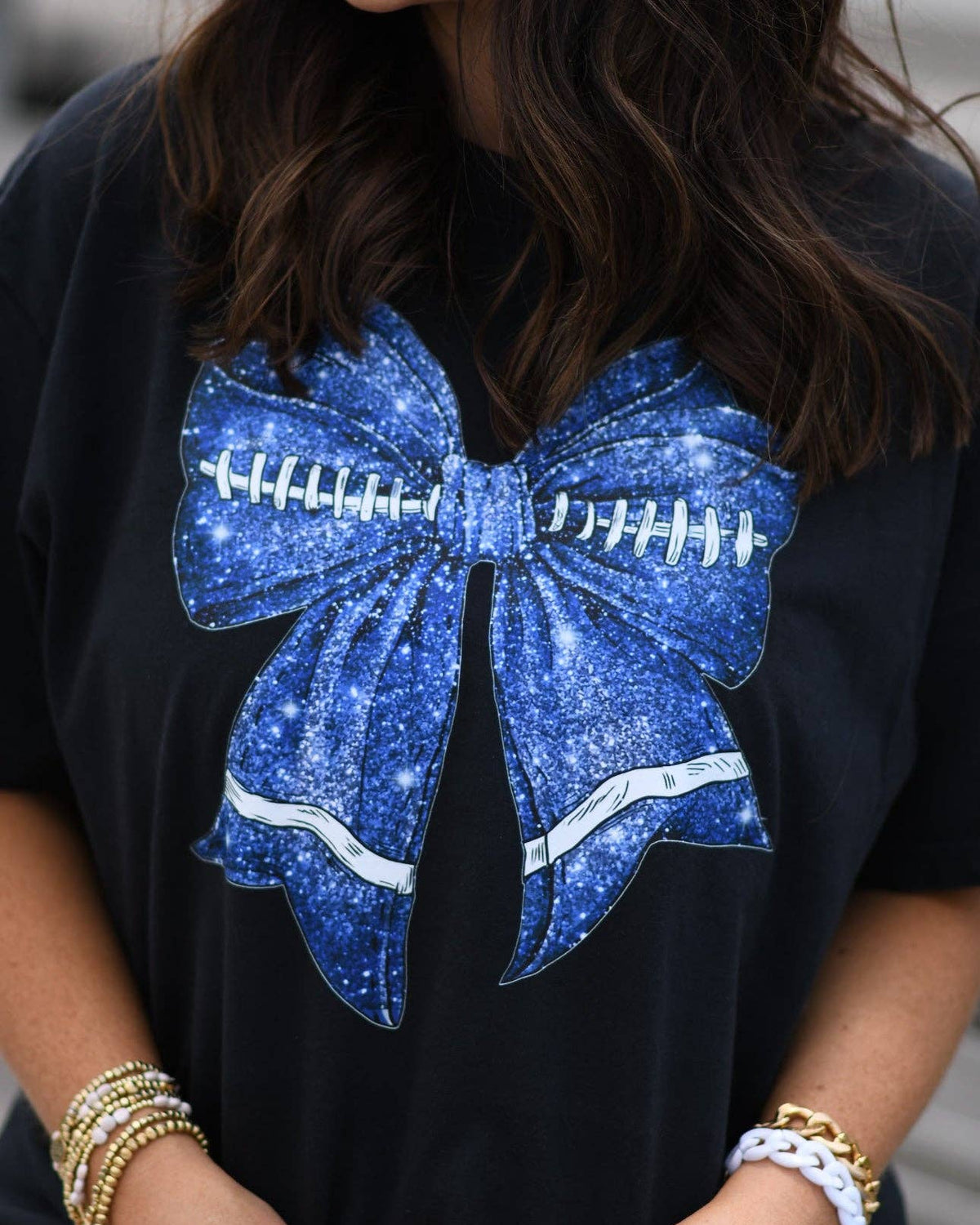a black Comfort Colors tee crafted on soft garment-dyed cotton with a DTF-printed blue faux glitter football bow graphic, crew neckline, and short sleeves in both youth and adult sizing