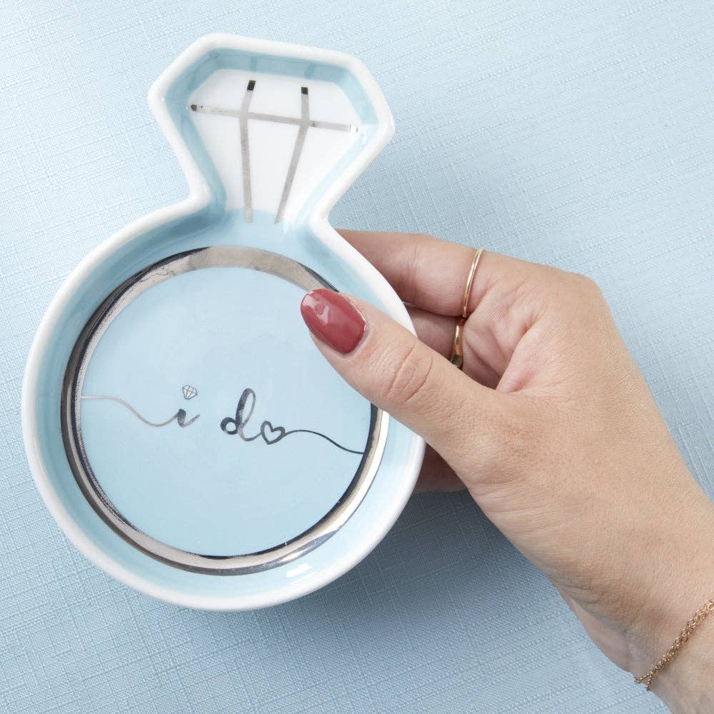 Hand holding a light blue ring holder with 'i do' text on a light blue background