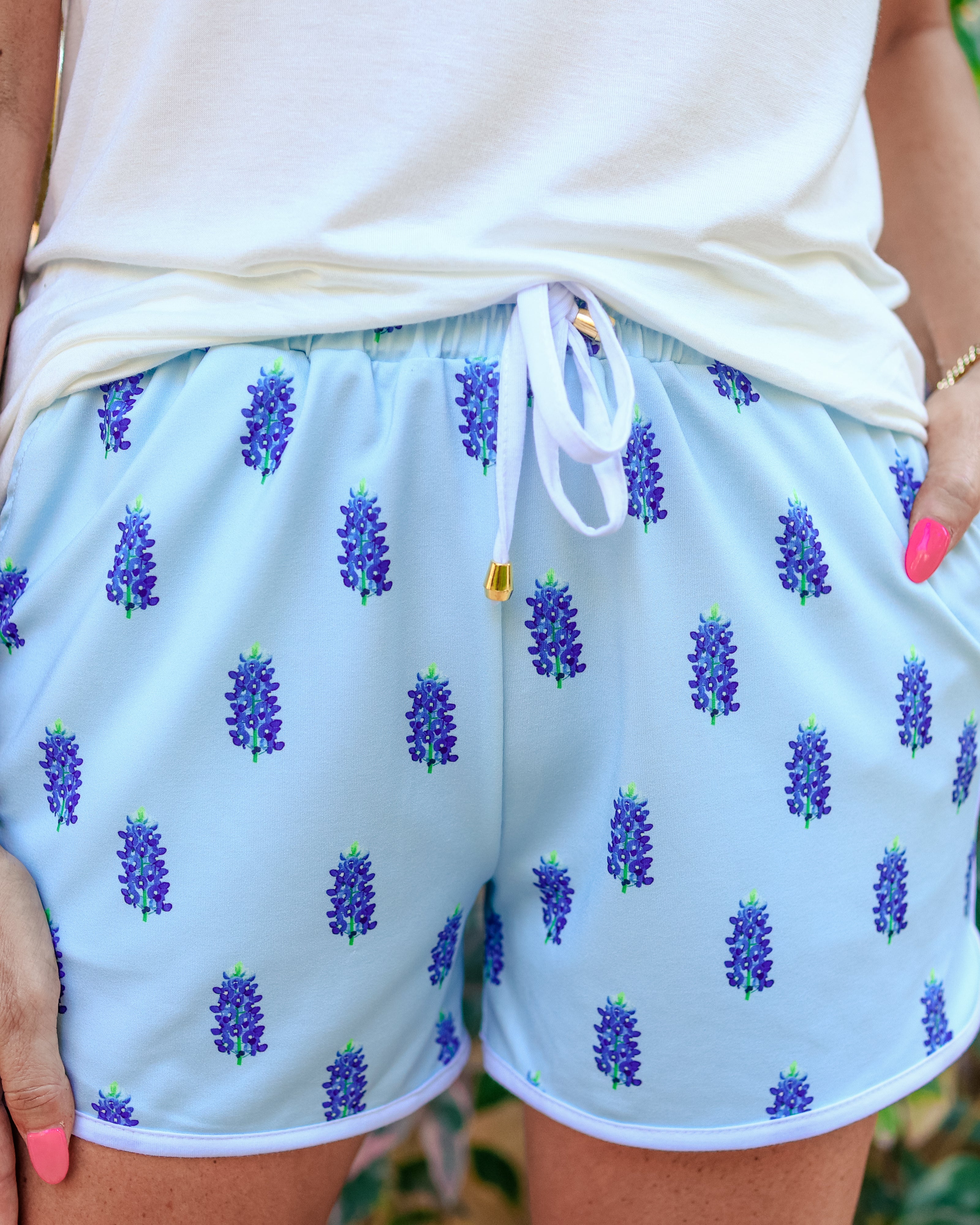 mid-rise relaxed fit activewear shorts with side pockets and a tie waist drawstring with gold bead accents, designed on a bluebonnet pattern print fabric, light blue base, and ivory trim