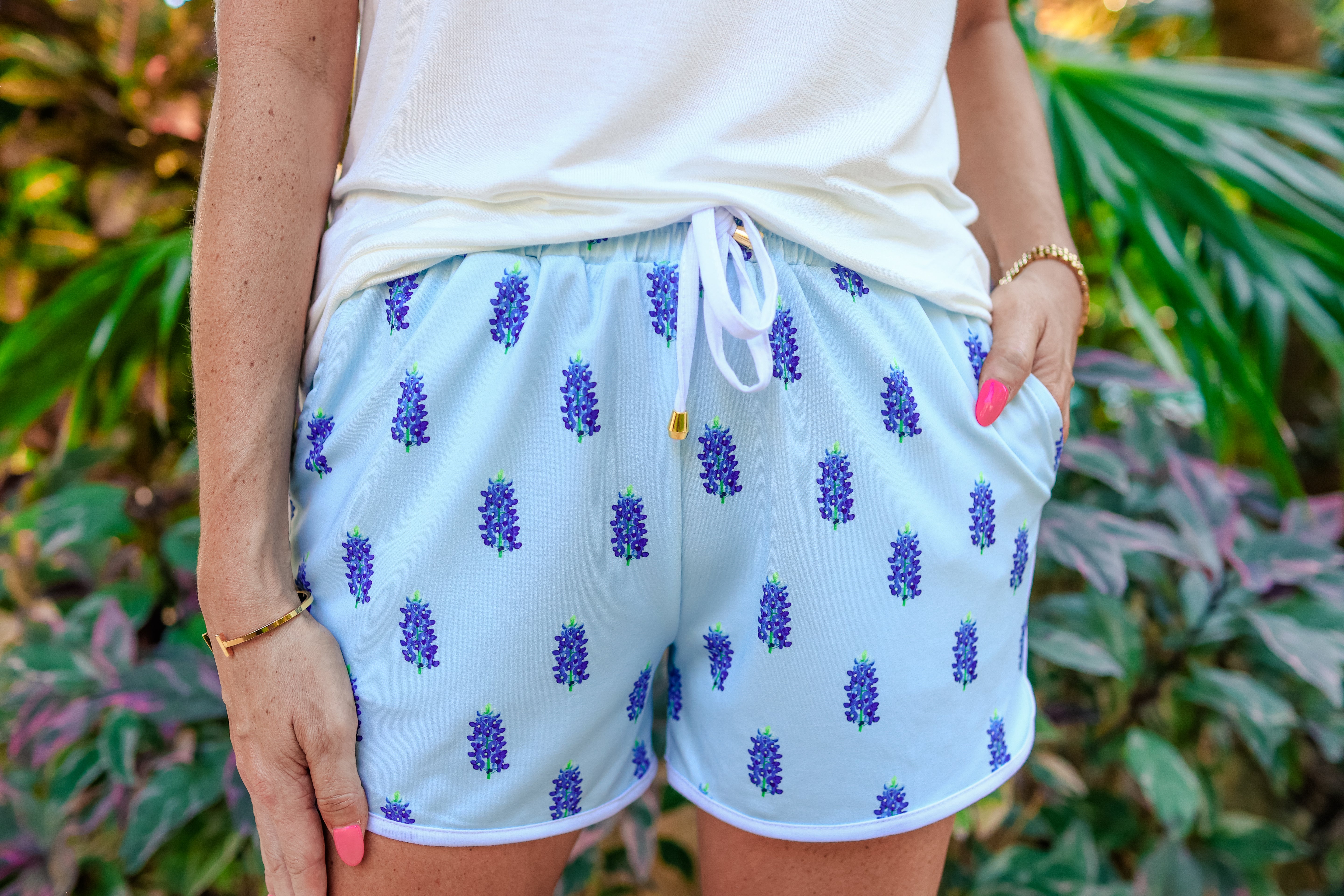 mid-rise relaxed fit activewear shorts with side pockets and a tie waist drawstring with gold bead accents, designed on a bluebonnet pattern print fabric, light blue base, and ivory trim