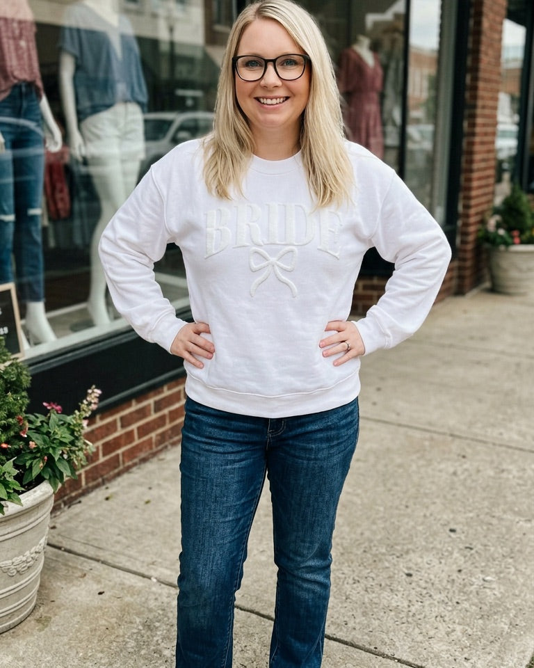 Woman wearing a white sweater and blue jeans standing on a sidewalk with a store in the background.