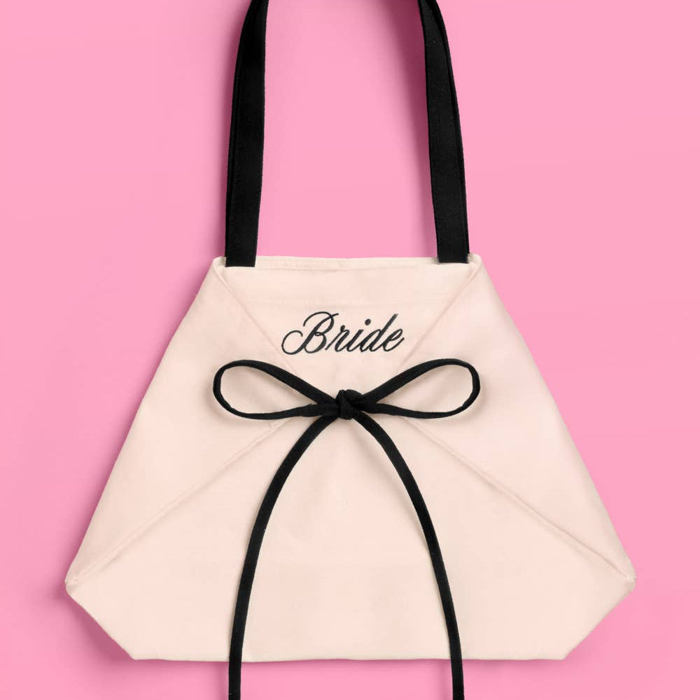 a roomy canvas tote with black embroidered 'Bride' script, black shoulder straps, and bow tie detail