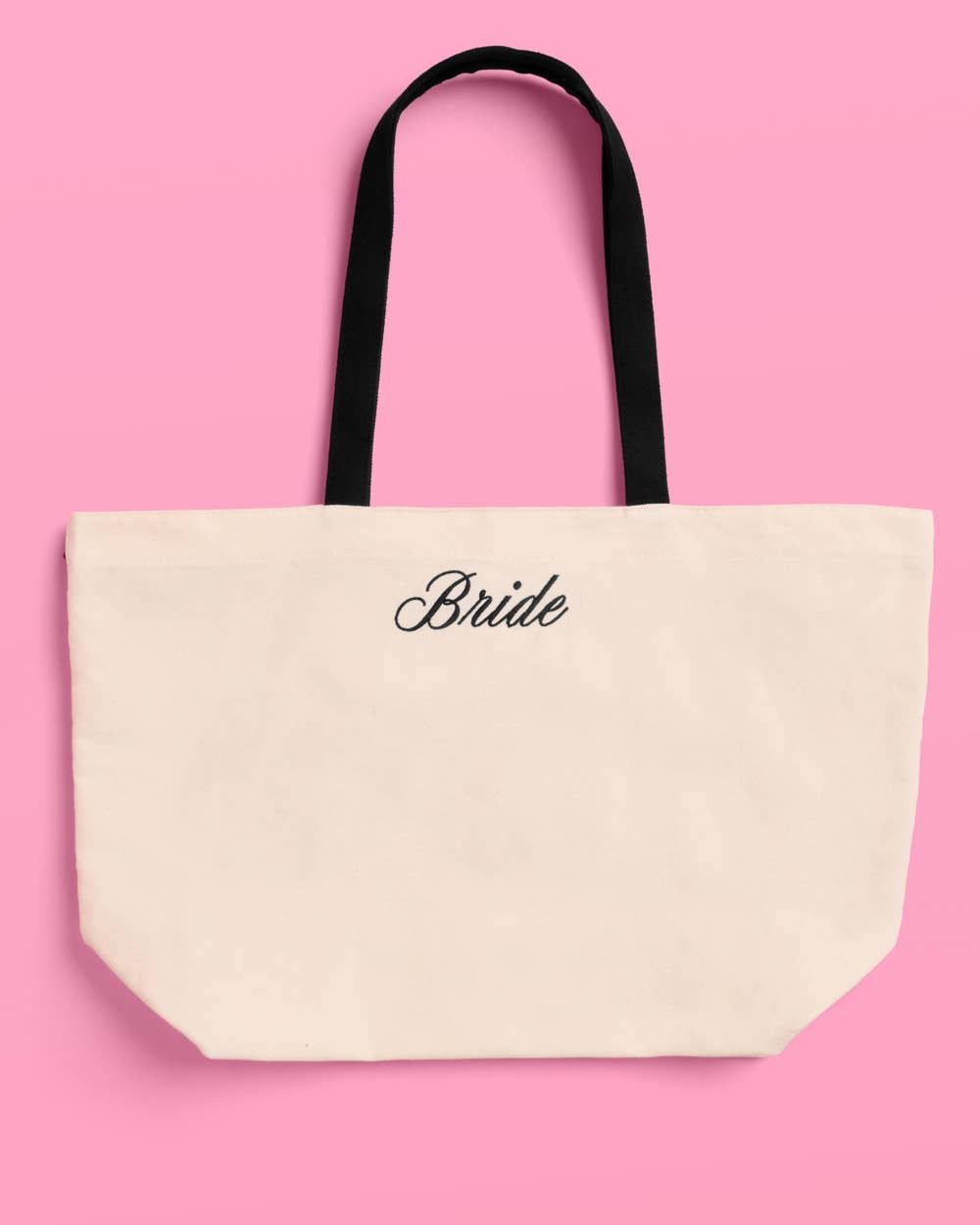 a roomy cream canvas tote with black embroidered 'Bride' script, black shoulder straps, and bow tie detail