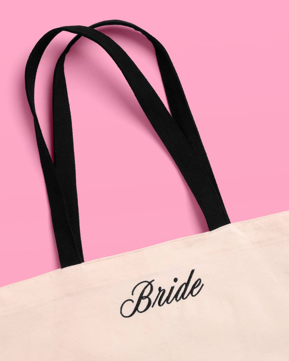 a roomy cream canvas tote with black embroidered 'Bride' script, black shoulder straps, and bow tie detail