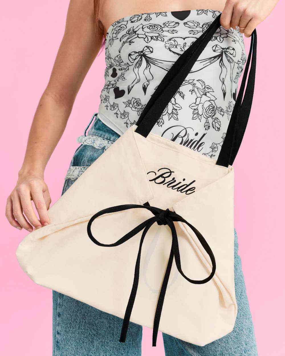 a roomy cream canvas tote with black embroidered 'Bride' script, black shoulder straps, and bow tie detail