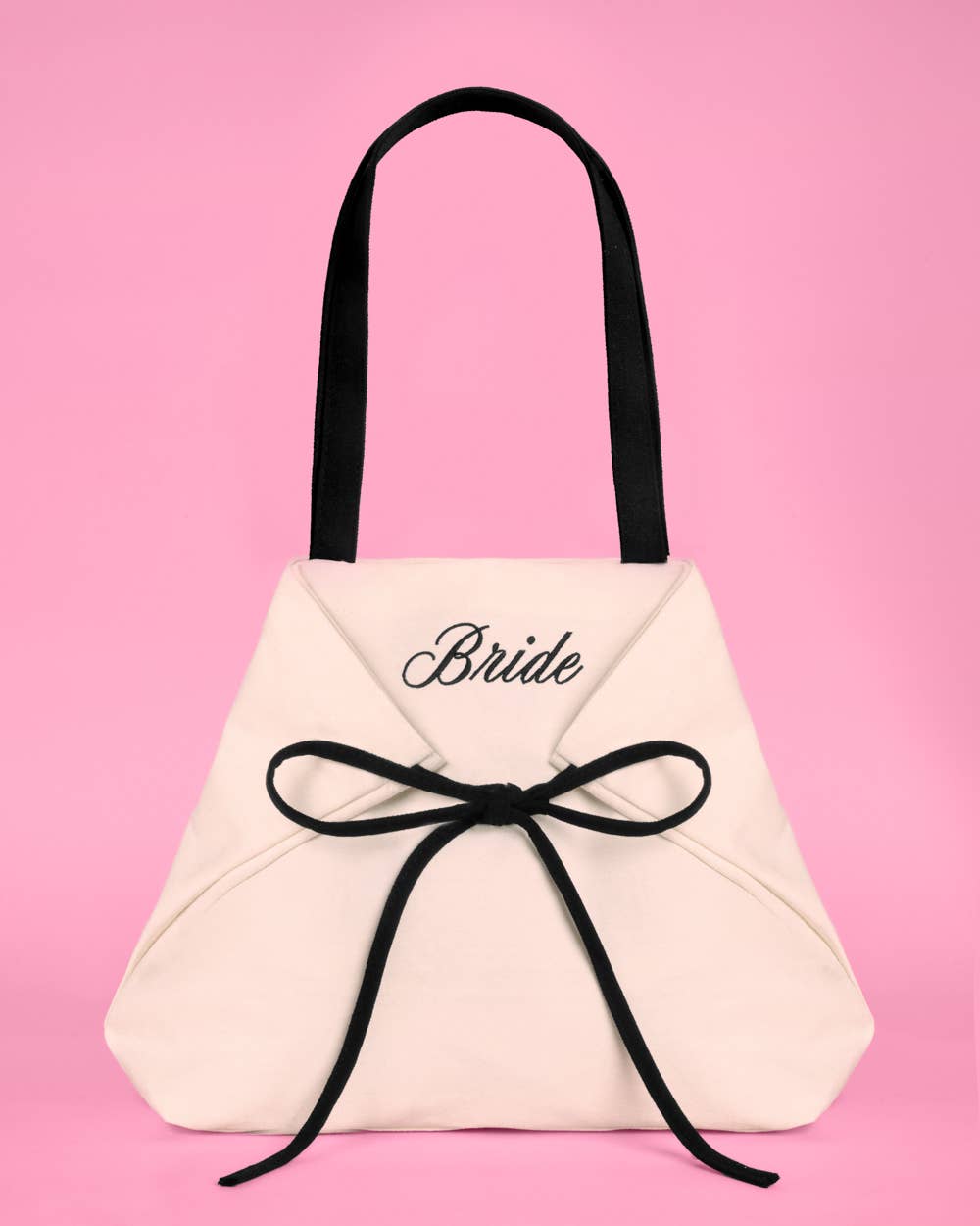 a roomy cream canvas tote with black embroidered 'Bride' script, black shoulder straps, and bow tie detail