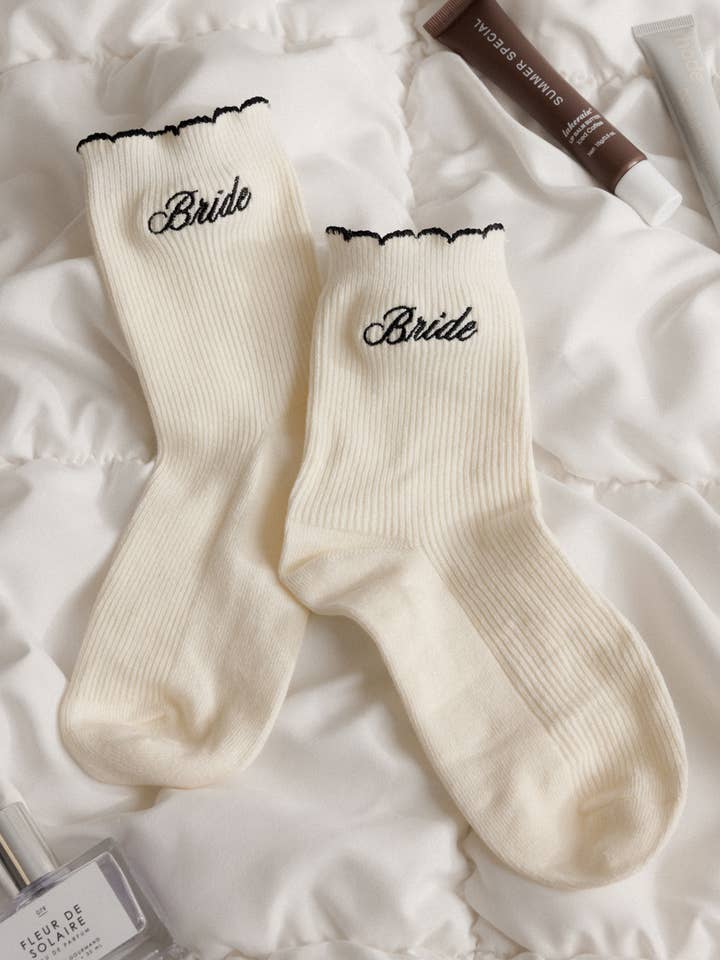 White socks with 'Bride' embroidery on a white surface with perfume bottles.