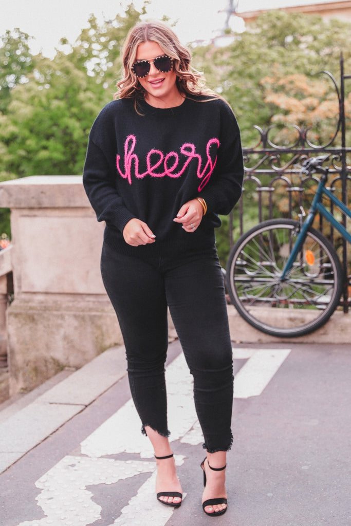 black long sleeve sweater adorned with 'cheers' in pink tinsel 