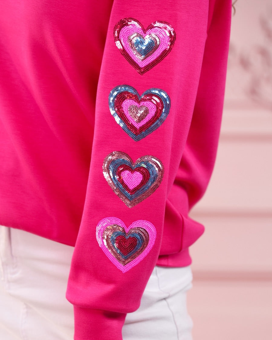 a hot pink long-sleeve pullover made from luxe material with a relaxed drop-shoulder silhouette, a classic crew neckline, and colorful sequin heart accents along the sleeves and chest