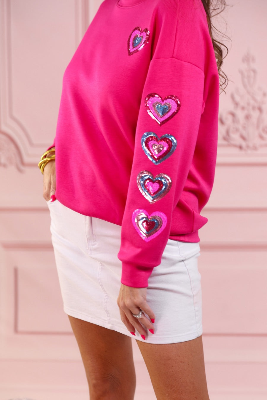 a hot pink long-sleeve pullover made from luxe material with a relaxed drop-shoulder silhouette, a classic crew neckline, and colorful sequin heart accents along the sleeves and chest