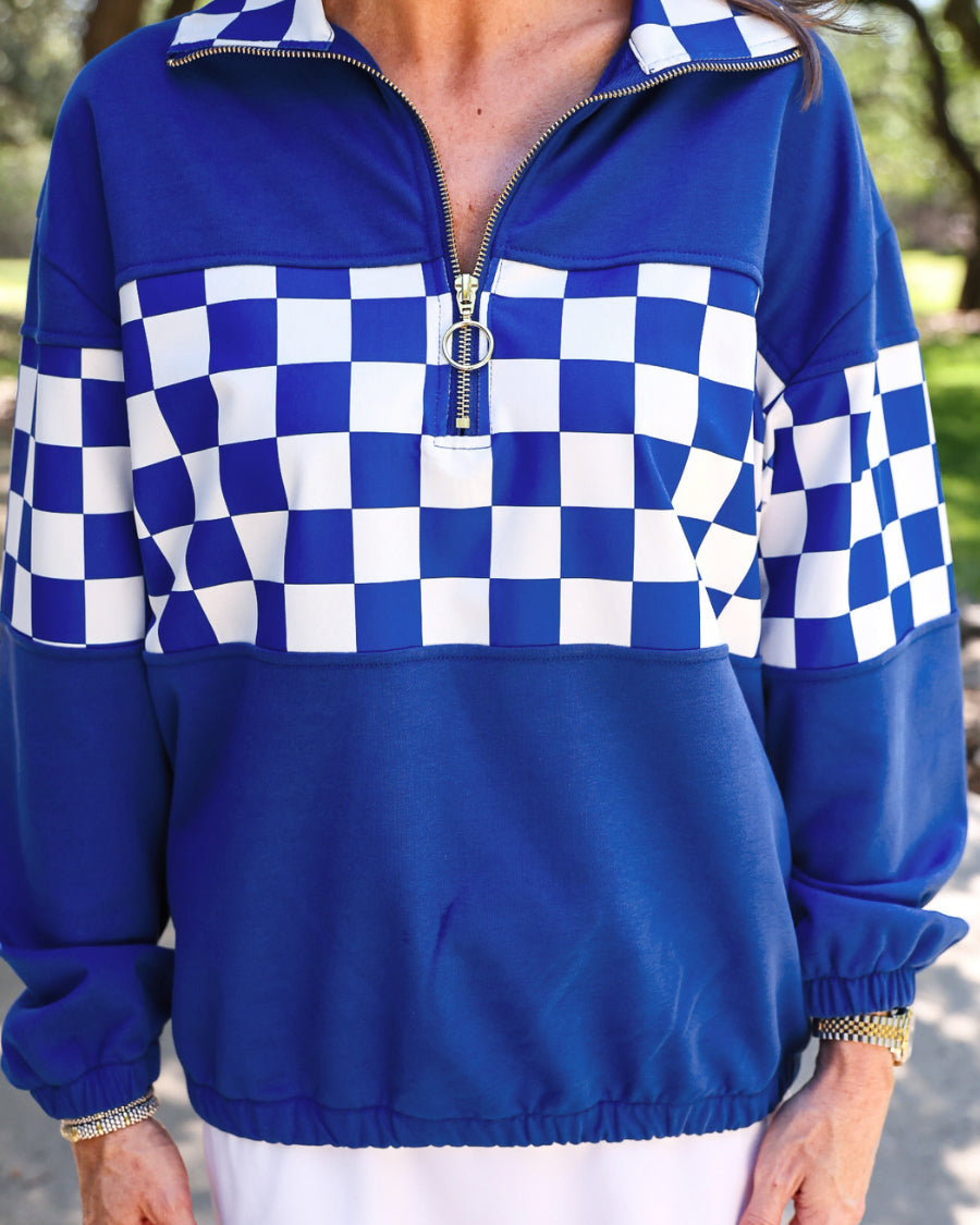a relaxed blue half-zip pullover with gold zipper detail, checkerboard contrast panels, long sleeves, elasticized cuffs, and a softly gathered bottom hem