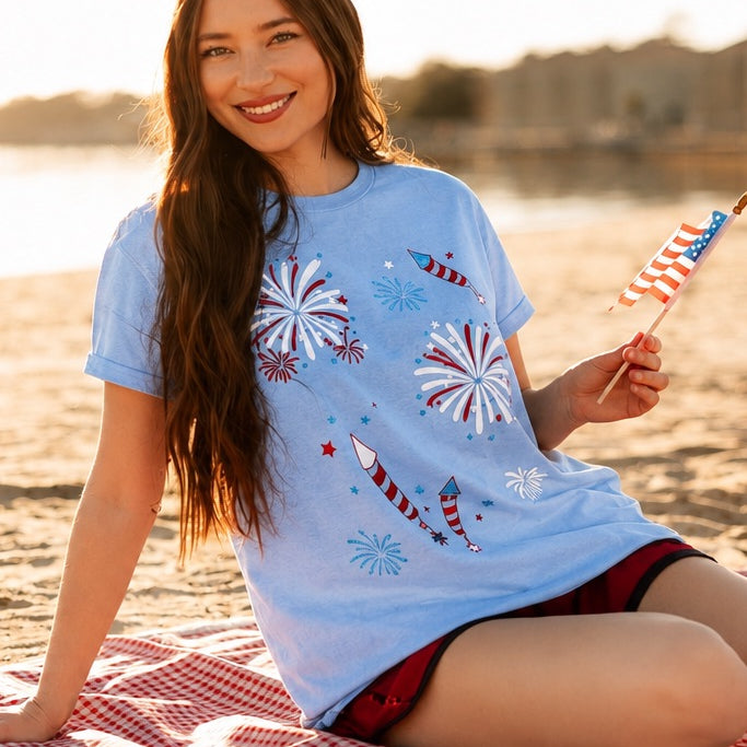 a relaxed blue crew neck tee with a screenprinted fireworks design (rockets, bursts, and starry details), short sleeves, standard t-shirt length, unlined construction, and soft cotton with light stretch