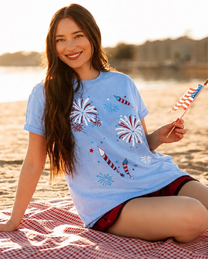 a relaxed blue crew neck tee with a screenprinted fireworks design (rockets, bursts, and starry details), short sleeves, standard t-shirt length, unlined construction, and soft cotton with light stretch