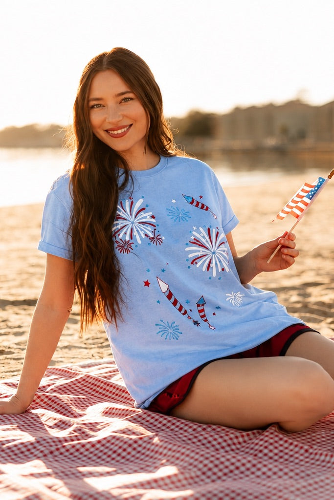 a relaxed blue crew neck tee with a screenprinted fireworks design (rockets, bursts, and starry details), short sleeves, standard t-shirt length, unlined construction, and soft cotton with light stretch