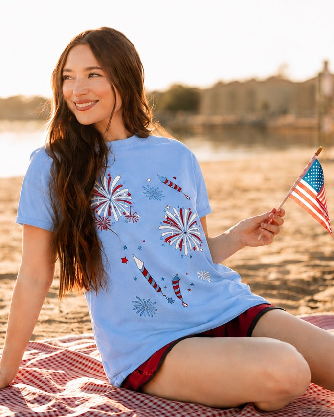 a relaxed blue crew neck tee with a screenprinted fireworks design (rockets, bursts, and starry details), short sleeves, standard t-shirt length, unlined construction, and soft cotton with light stretch