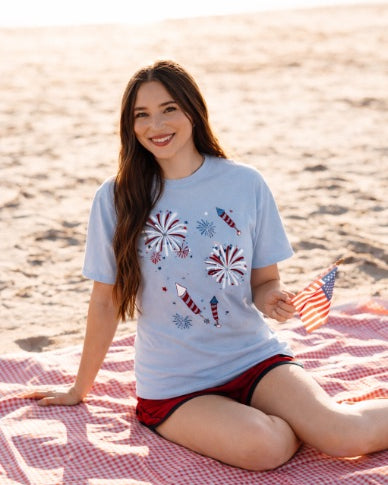 a relaxed blue crew neck tee with a screenprinted fireworks design (rockets, bursts, and starry details), short sleeves, standard t-shirt length, unlined construction, and soft cotton with light stretch