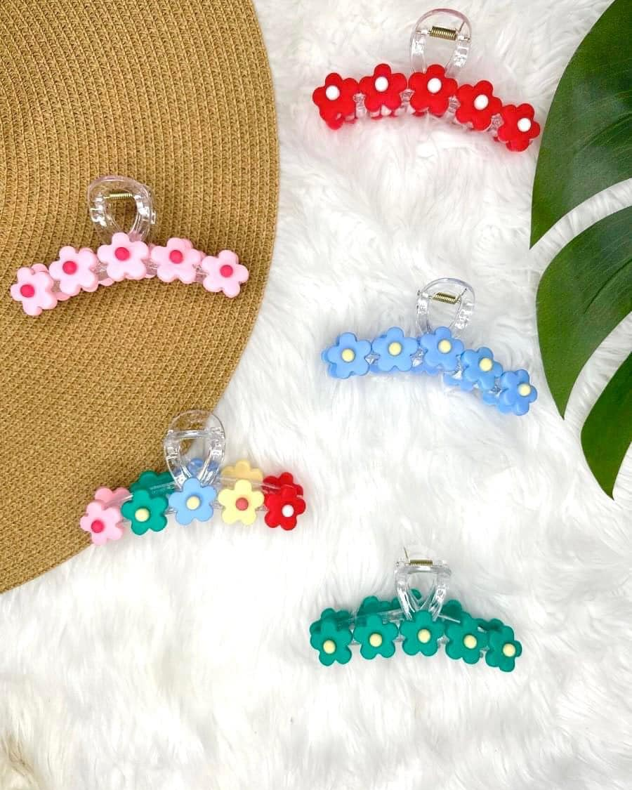 Colorful daily-inspired bloom hair clips on a fluffy white surface with a straw hat and green leaf in the background.