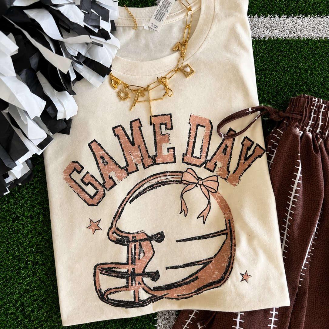 a heather natural Bella+Canvas unisex soft tee with a DTF-printed vintage-inspired football helmet, bow accent, arched 'GAME DAY' varsity lettering, star details, crew neckline, short sleeves, and a relaxed fit in both youth and adult sizing