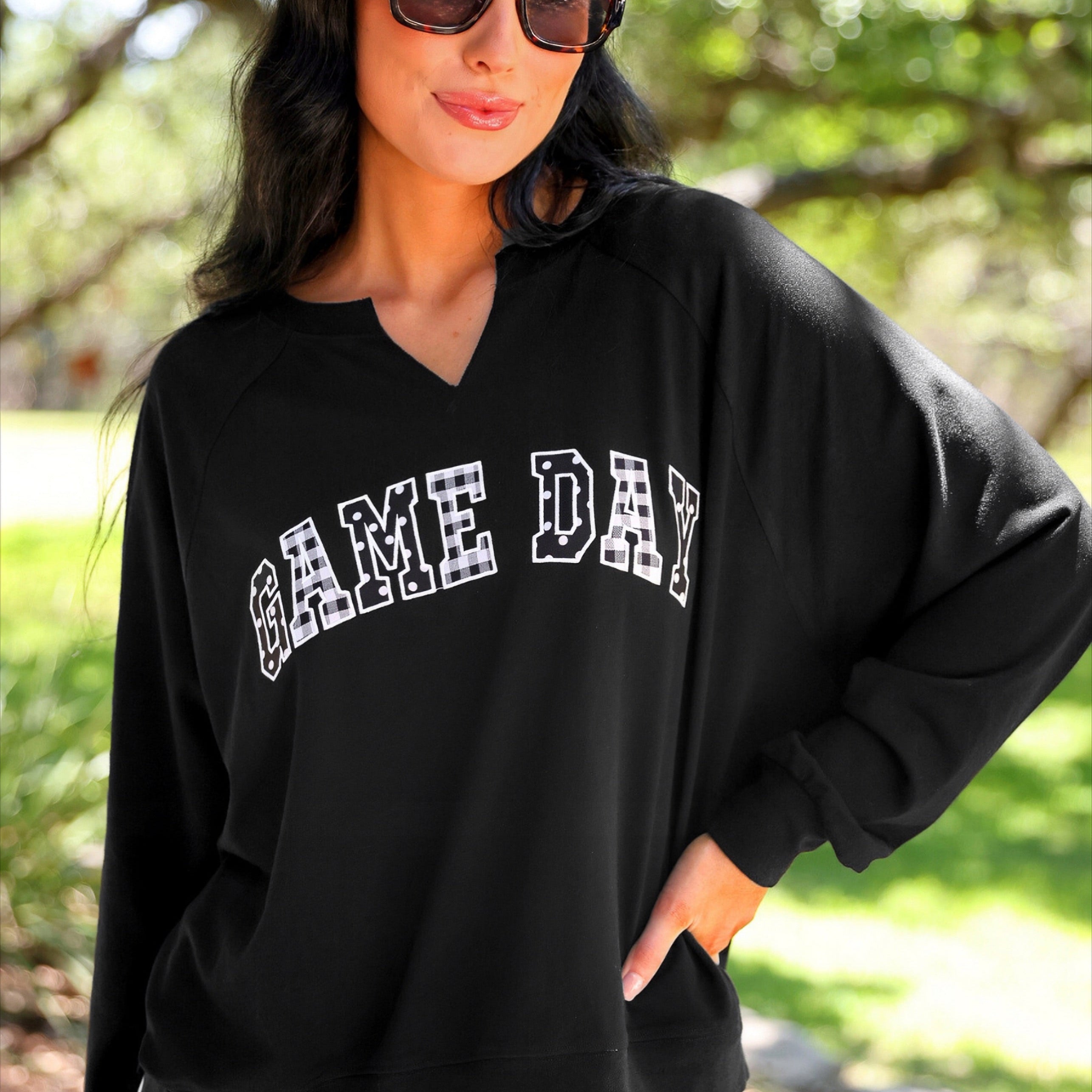 a relaxed black French terry pullover with a V-neckline, mixed polka dot and gingham 'GAME DAY' lettering, long sleeves, banded cuffs, and side slit hems on a cotton fabric
