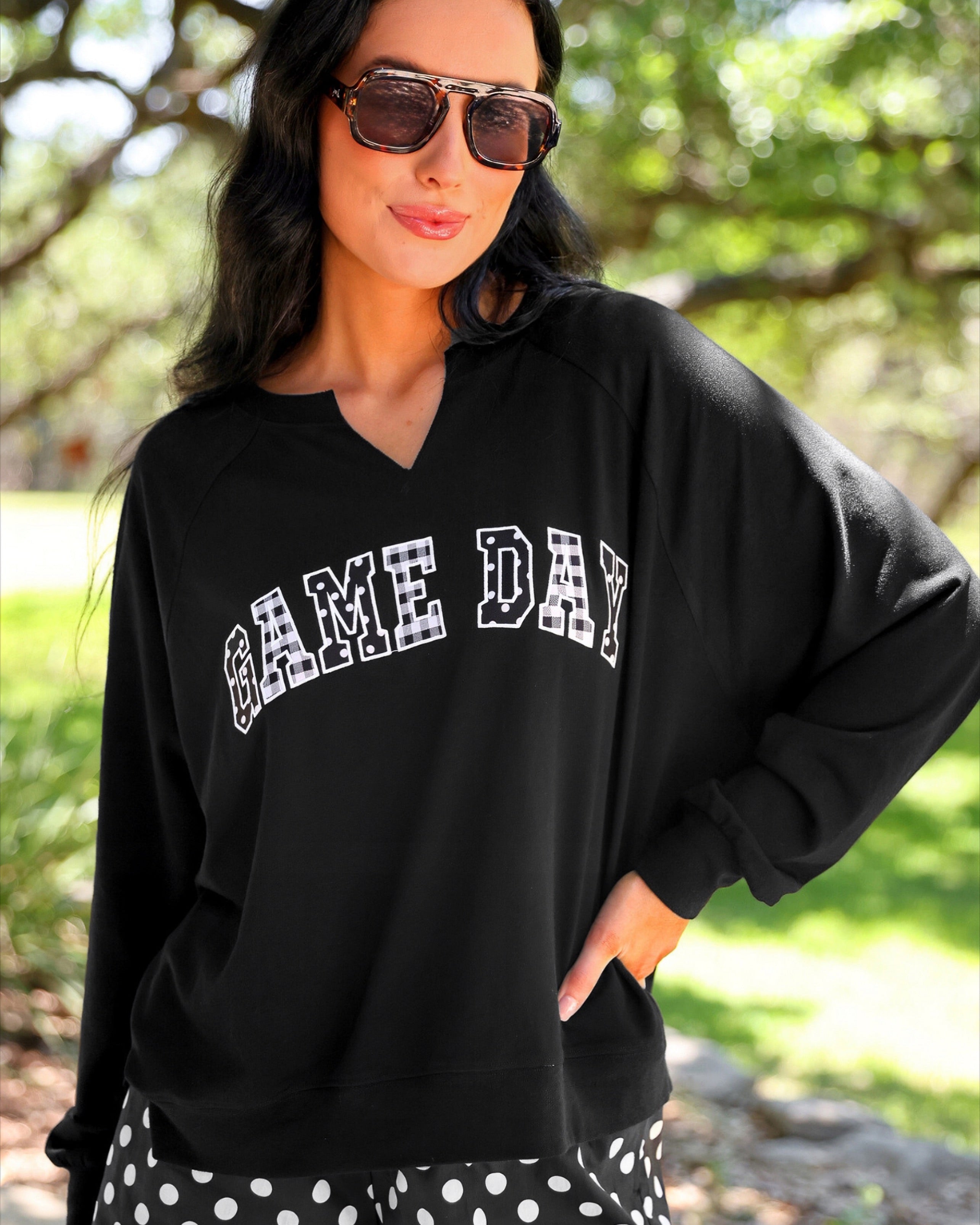 a relaxed black French terry pullover with a V-neckline, mixed polka dot and gingham 'GAME DAY' lettering, long sleeves, banded cuffs, and side slit hems on a cotton fabric