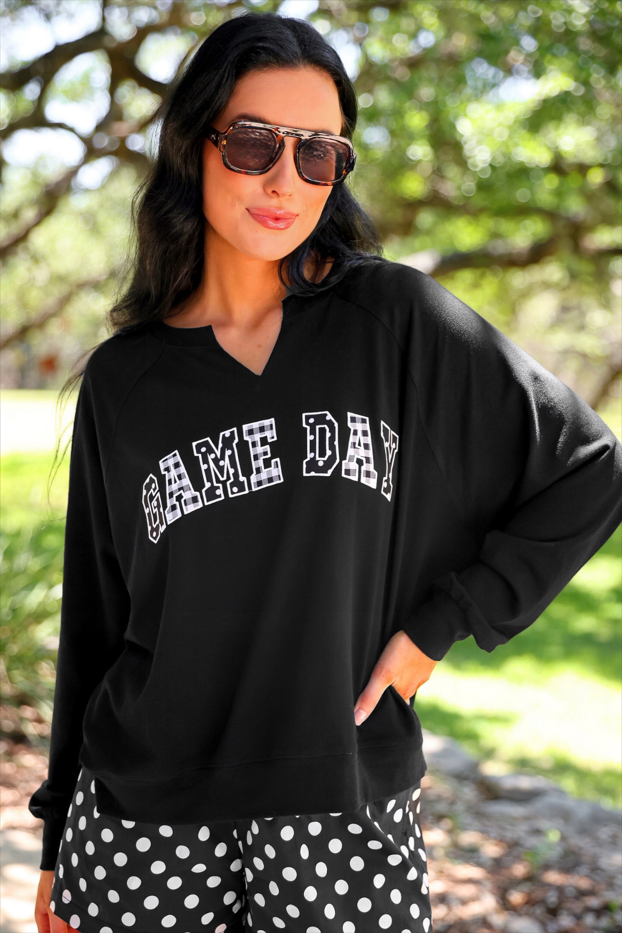 a relaxed black French terry pullover with a V-neckline, mixed polka dot and gingham 'GAME DAY' lettering, long sleeves, banded cuffs, and side slit hems on a cotton fabric
