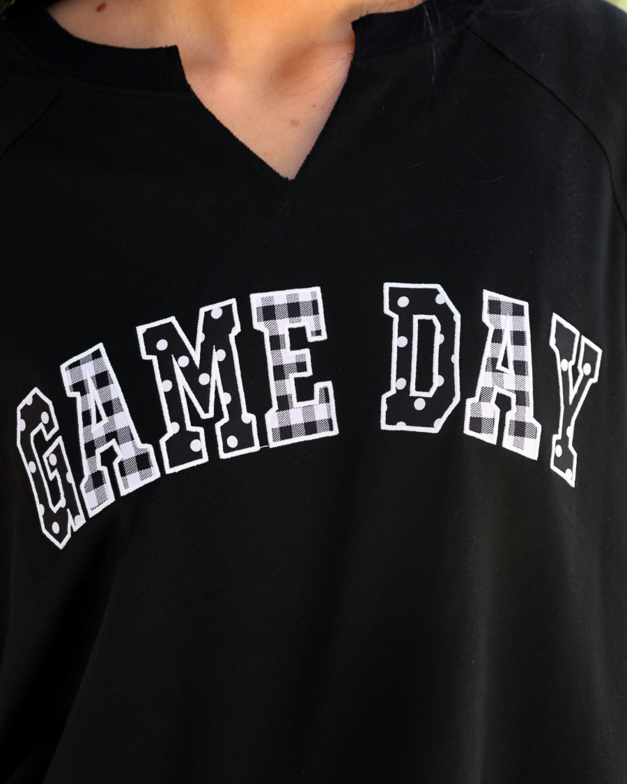 a relaxed black French terry pullover with a V-neckline, mixed polka dot and gingham 'GAME DAY' lettering, long sleeves, banded cuffs, and side slit hems on a cotton fabric