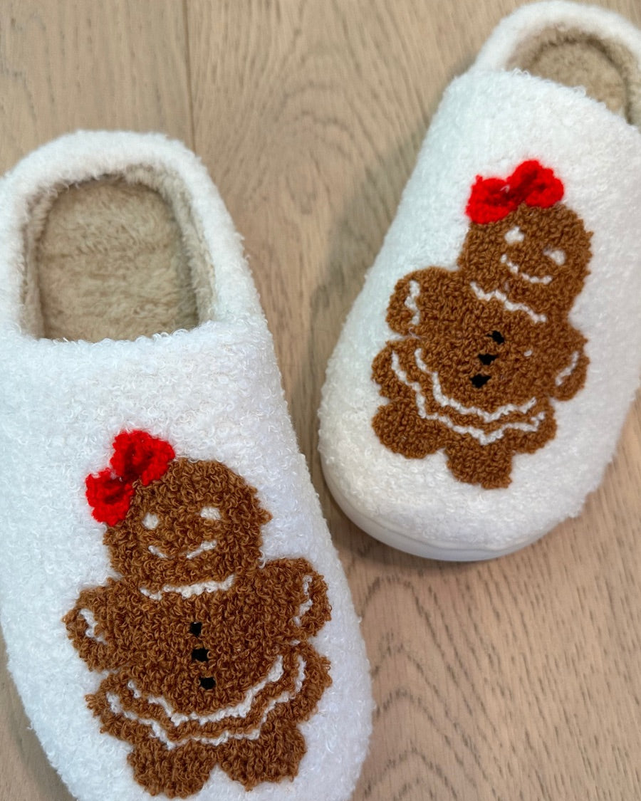 white plush slippers crafted from sherpa style material adorned with an adorable chenille gingerbread girl topped with a festive red bow, a soft interior lining, and easy slip-on silhouette