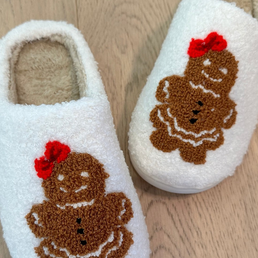 white plush slippers crafted from sherpa style material adorned with an adorable chenille gingerbread girl topped with a festive red bow, a soft interior lining, and easy slip-on silhouette