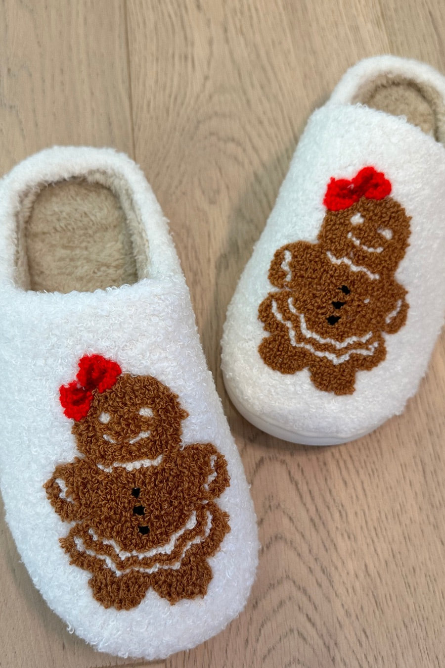 white plush slippers crafted from sherpa style material adorned with an adorable chenille gingerbread girl topped with a festive red bow, a soft interior lining, and easy slip-on silhouette