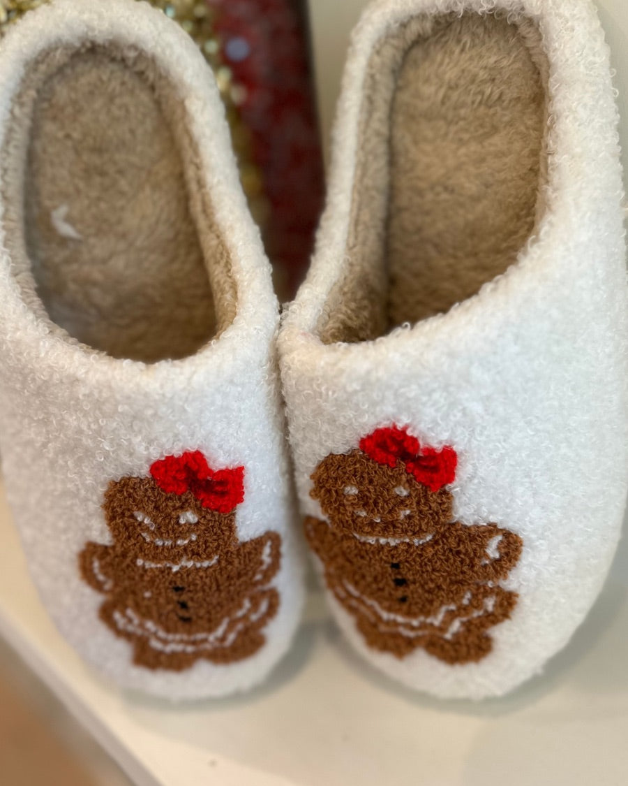 white plush slippers crafted from sherpa style material adorned with an adorable chenille gingerbread girl topped with a festive red bow, a soft interior lining, and easy slip-on silhouette