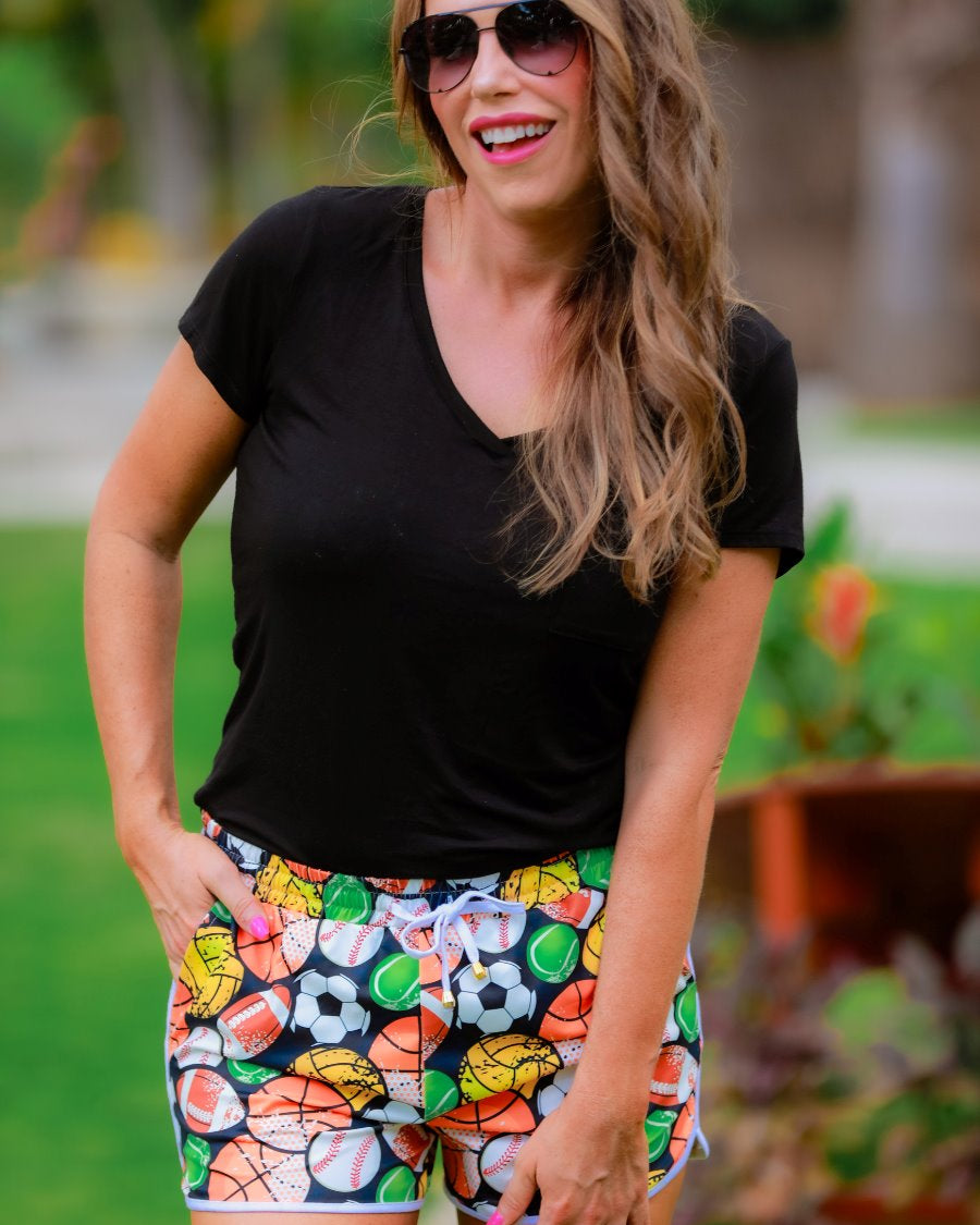 mid-rise relaxed fit activewear shorts with side pockets and a tie waist drawstring with gold bead accents, designed on a multi-sport pattern print fabric, black base, and ivory trim