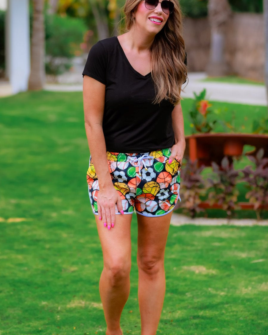 mid-rise relaxed fit activewear shorts with side pockets and a tie waist drawstring with gold bead accents, designed on a multi-sport pattern print fabric, black base, and ivory trim