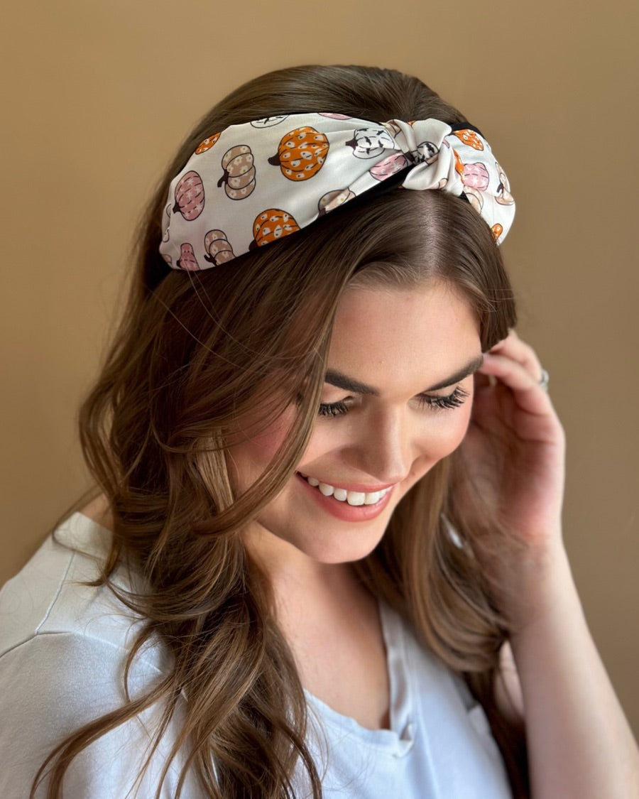 a twisted center top knot headband crafted from soft, lightweight material with a multicolor jack-o-lantern pattern on a ivory base