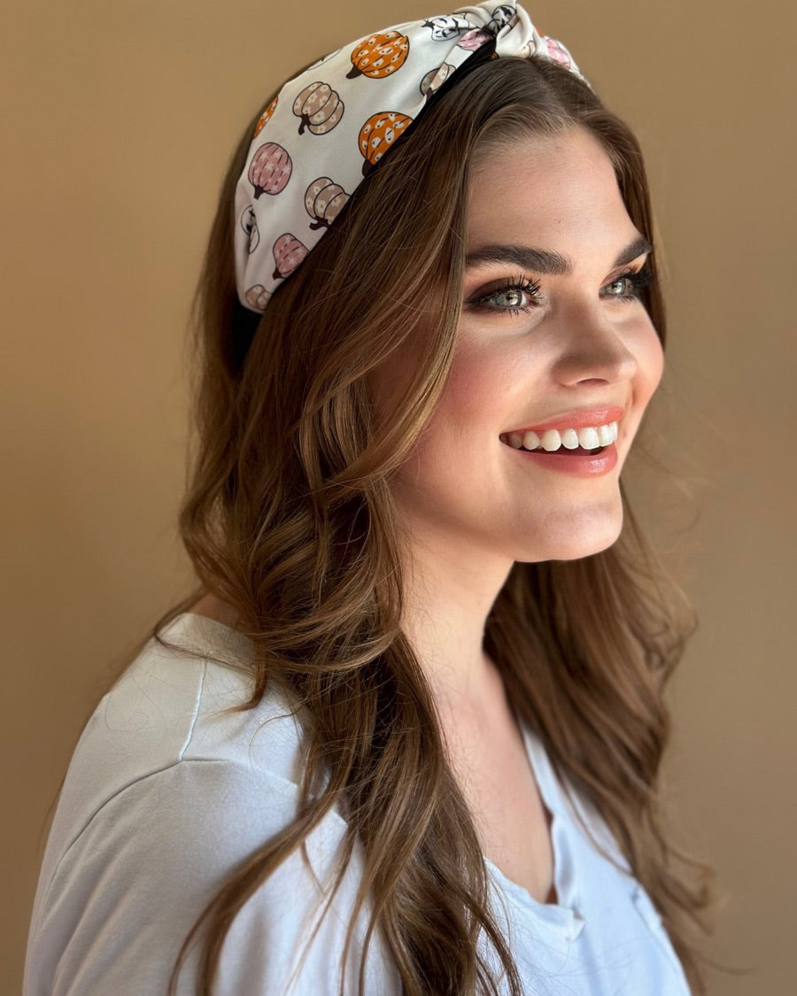 a twisted center top knot headband crafted from soft, lightweight material with a multicolor jack-o-lantern pattern on a ivory base