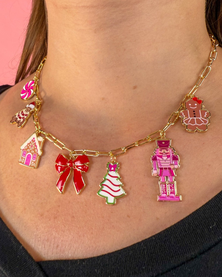 an adjustable gold link paperclip-style necklace adorned with whimsical enamel charms (a bold red bow, Christmas bear, Christmas tree, gingerbread girl, gingerbread house, pink nutcracker, and a pink peppermint)