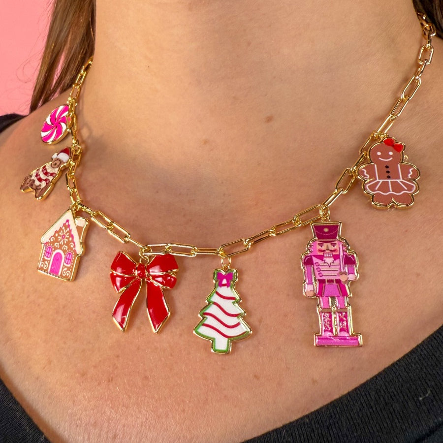 an adjustable gold link paperclip-style necklace adorned with whimsical enamel charms (a bold red bow, Christmas bear, Christmas tree, gingerbread girl, gingerbread house, pink nutcracker, and a pink peppermint)