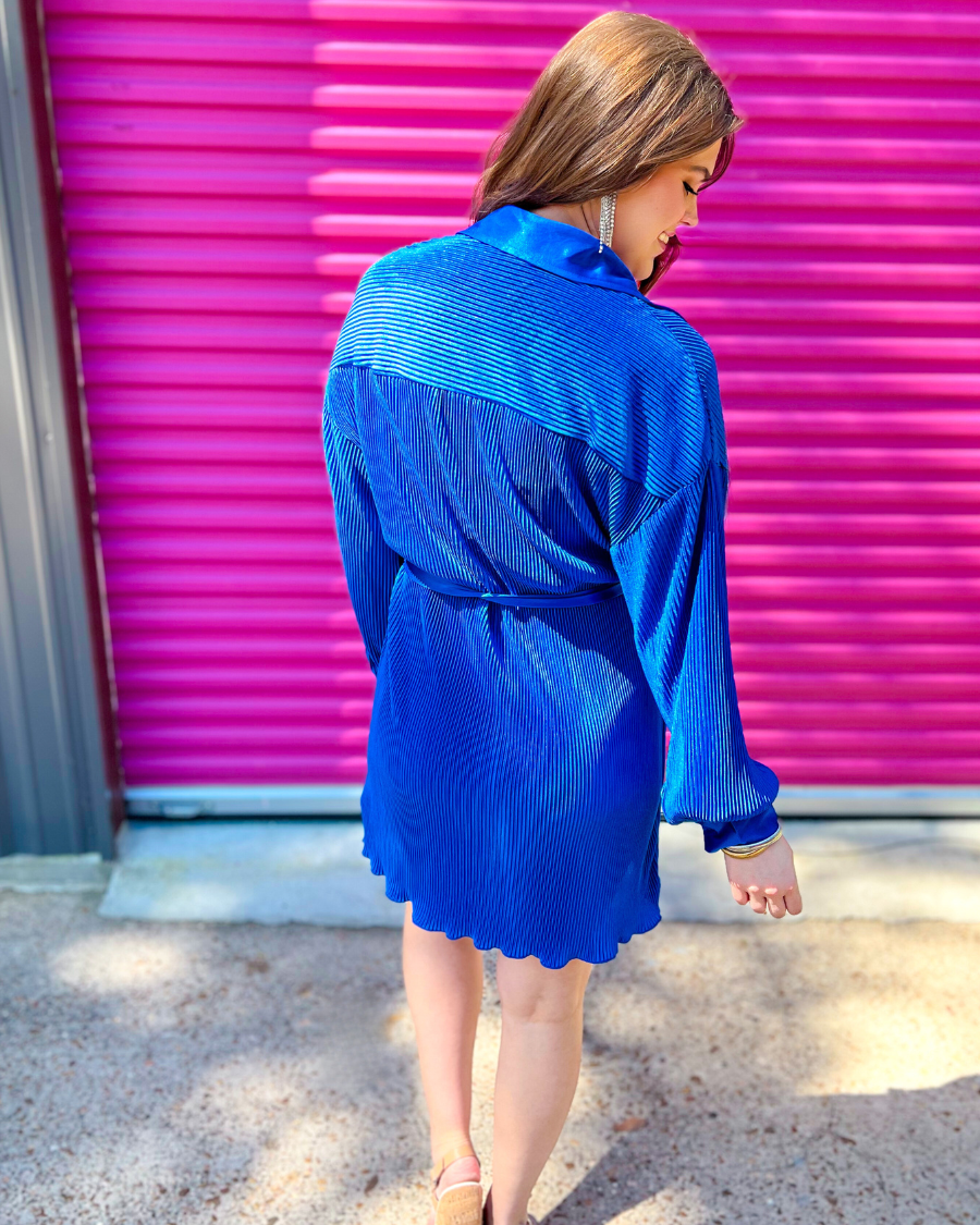 a royal blue pleated shirt dress with a classic collar, functional button front, removable waist sash, drop shoulder long sleeves, satin cuffs, back yoke seam, accordion texture, and flowy mini-length silhouette