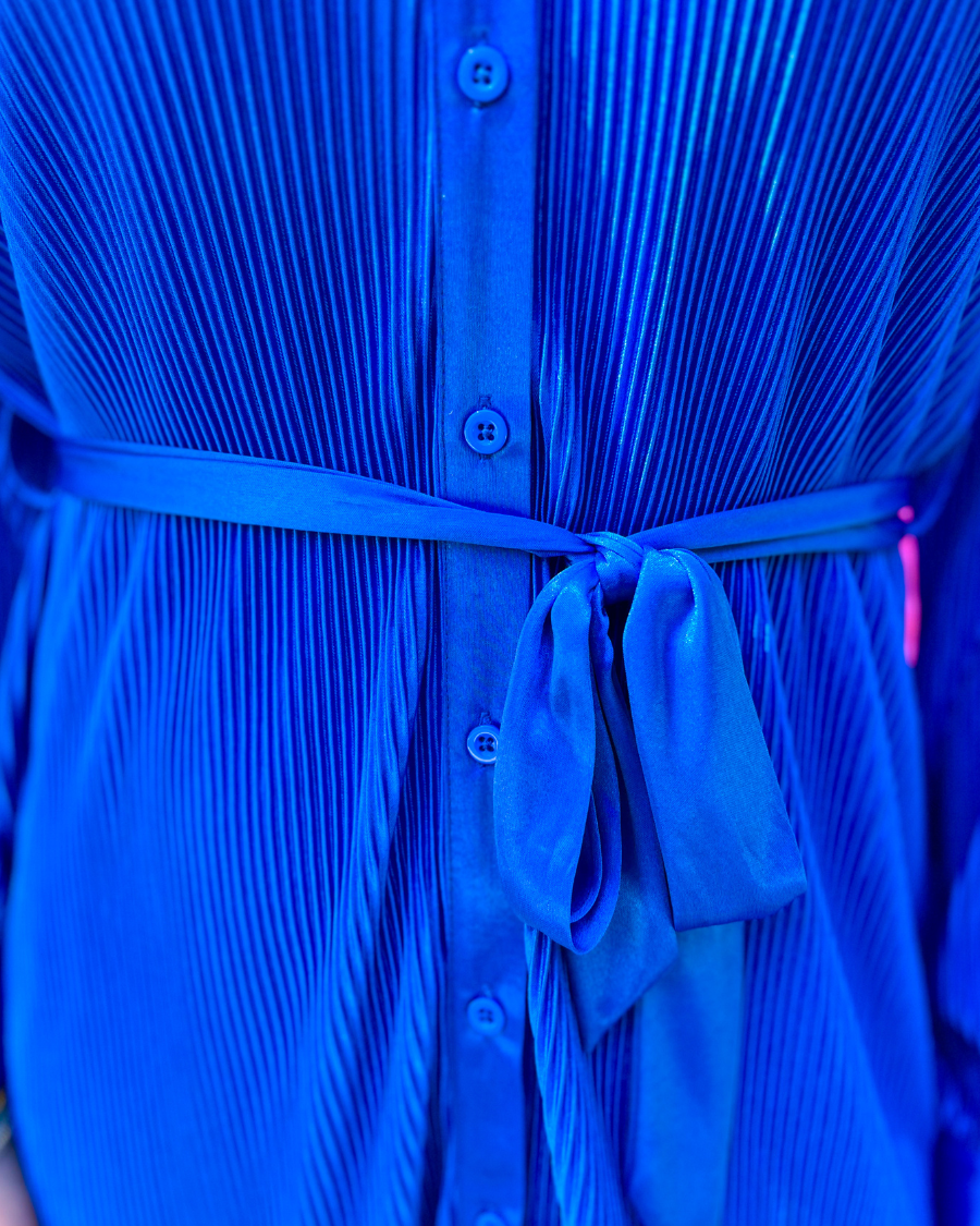 a royal blue pleated shirt dress with a classic collar, functional button front, removable waist sash, drop shoulder long sleeves, satin cuffs, back yoke seam, accordion texture, and flowy mini-length silhouette