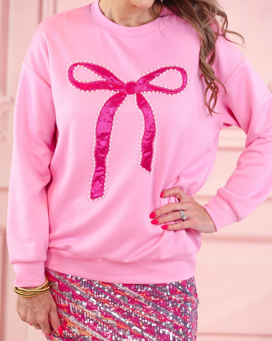 a light pink long-sleeve pullover made from luxe material with drop shoulder, a crew neckline, and a foil bow outlined in delicate pearls