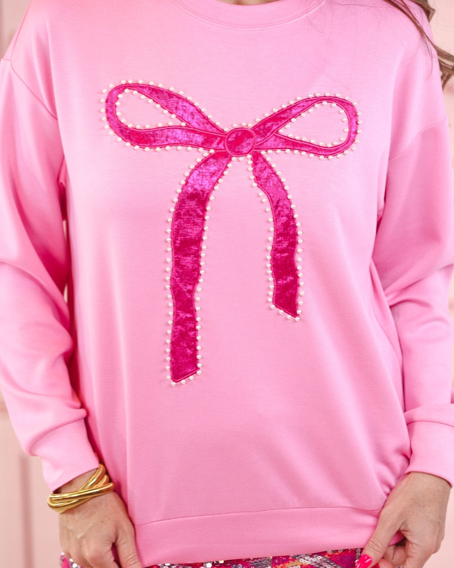 a light pink long-sleeve pullover made from luxe material with drop shoulder, a crew neckline, and a foil bow outlined in delicate pearls