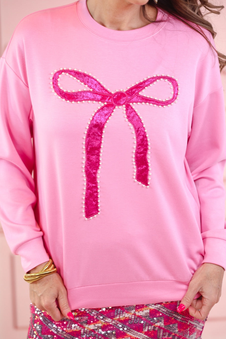 a light pink long-sleeve pullover made from luxe material with drop shoulder, a crew neckline, and a foil bow outlined in delicate pearls