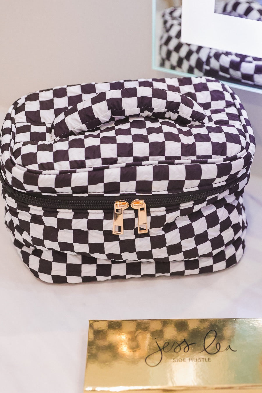 a structured bag with a spacious interior, a secure double zip around closure, a top handle, and gold accents on a quilted body in checkerboard checkered (black/white)